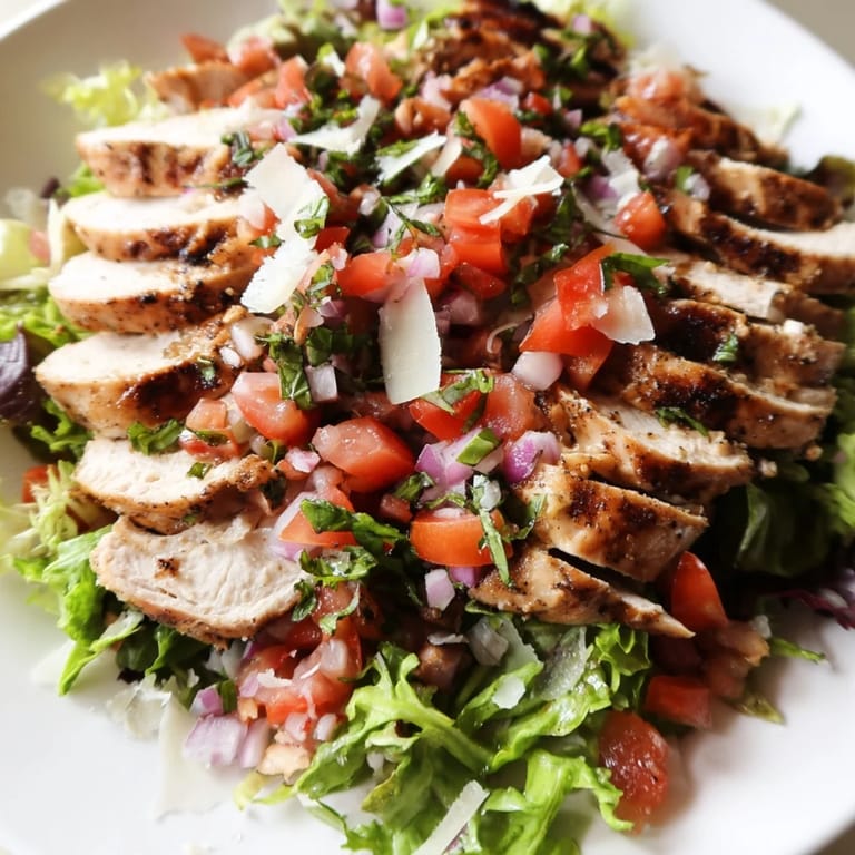 A plated Bruschetta Chicken Salad showcases sliced grilled chicken breast, a chunky tomato and garlic bruschetta topping, and shaved Parmesan over a bed of mixed greens.