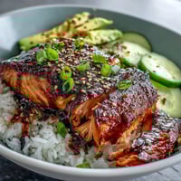Vibrant teriyaki salmon bowl with glazed fillets, jasmine rice, avocado, cucumber, and sesame seeds, served fresh for a quick summer dinner.